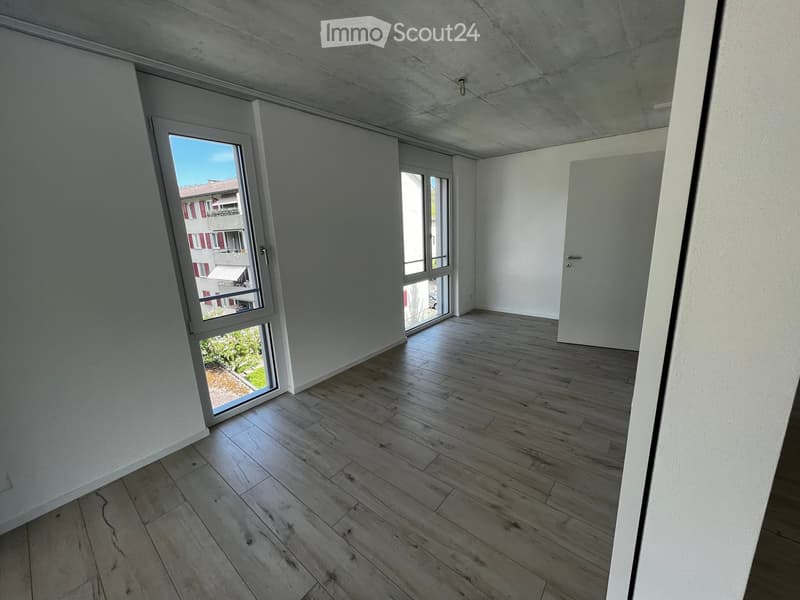 Empty room with light wooden flooring, white walls, and large windows offering a view of the outside. No furniture or equipment is present.