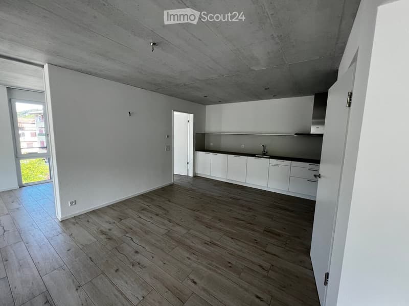 Empty room with white walls, wooden flooring, and a small kitchen area with white cabinets and a sink.