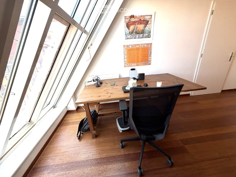 A modern, well-lit room with wooden flooring, large windows, a desk, ergonomic chair, laptop, and decorative items on the wall.