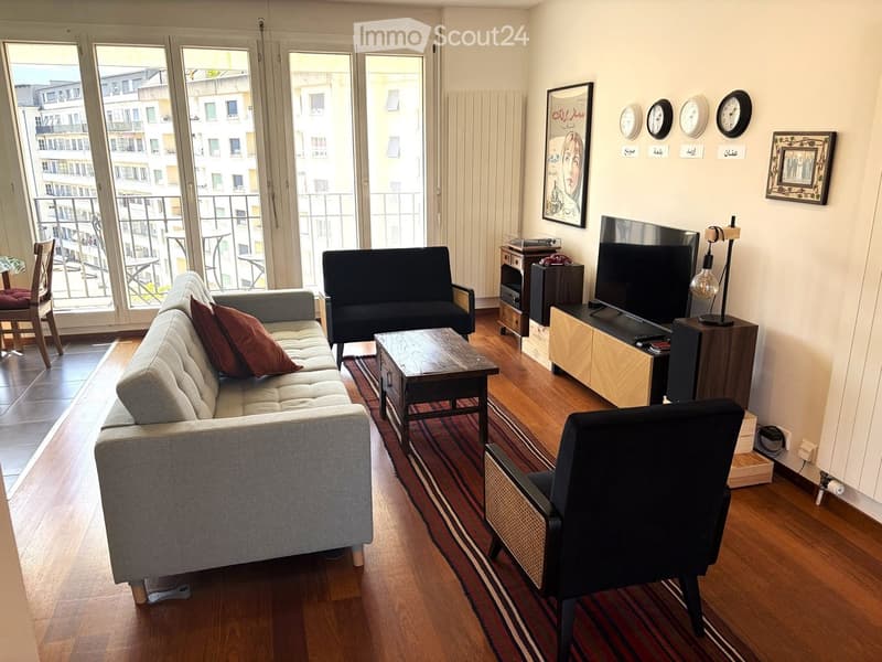 Modern living room with large windows offering a city view, featuring a grey sofa, coffee table, wooden floor, and a television on a wooden stand.