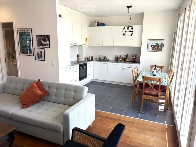 A modern, open-concept living and dining area with a grey sofa, wooden dining table, and white kitchen cabinets.