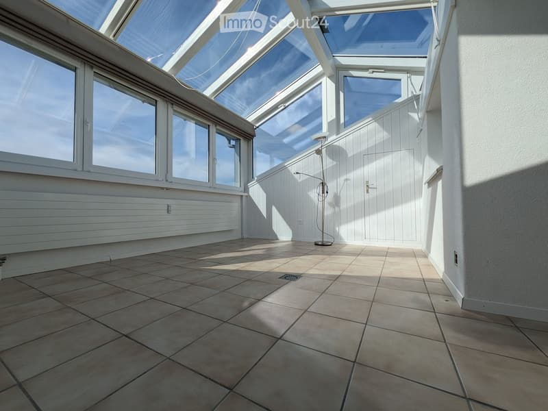 Bright, sunlit balcony with large glass roof panels, tiled floor, white wooden paneling, and a single floor lamp.