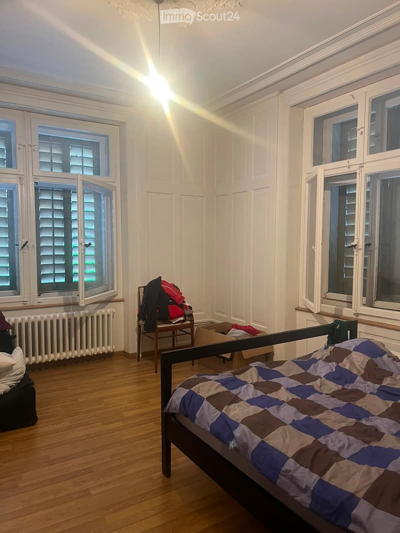 Bright bedroom with wooden flooring, large windows, a bed with a patterned quilt, a chair with clothes, and a radiator.