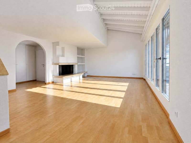 Empty room with light wooden flooring, white walls, and large windows allowing natural light. Features include a fireplace with built-in shelving and an arched doorway leading to another area.