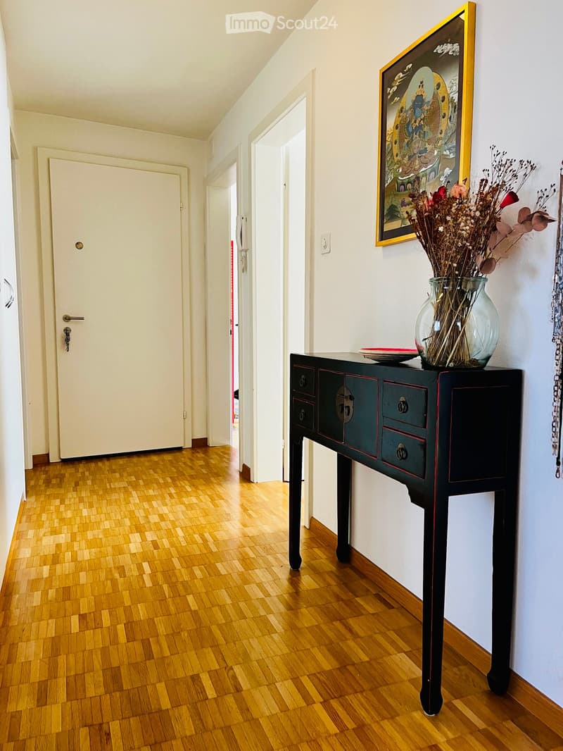 Hallway with wooden flooring, a small black table with a vase of dried flowers and a decorative plate, and a framed picture on the wall.
