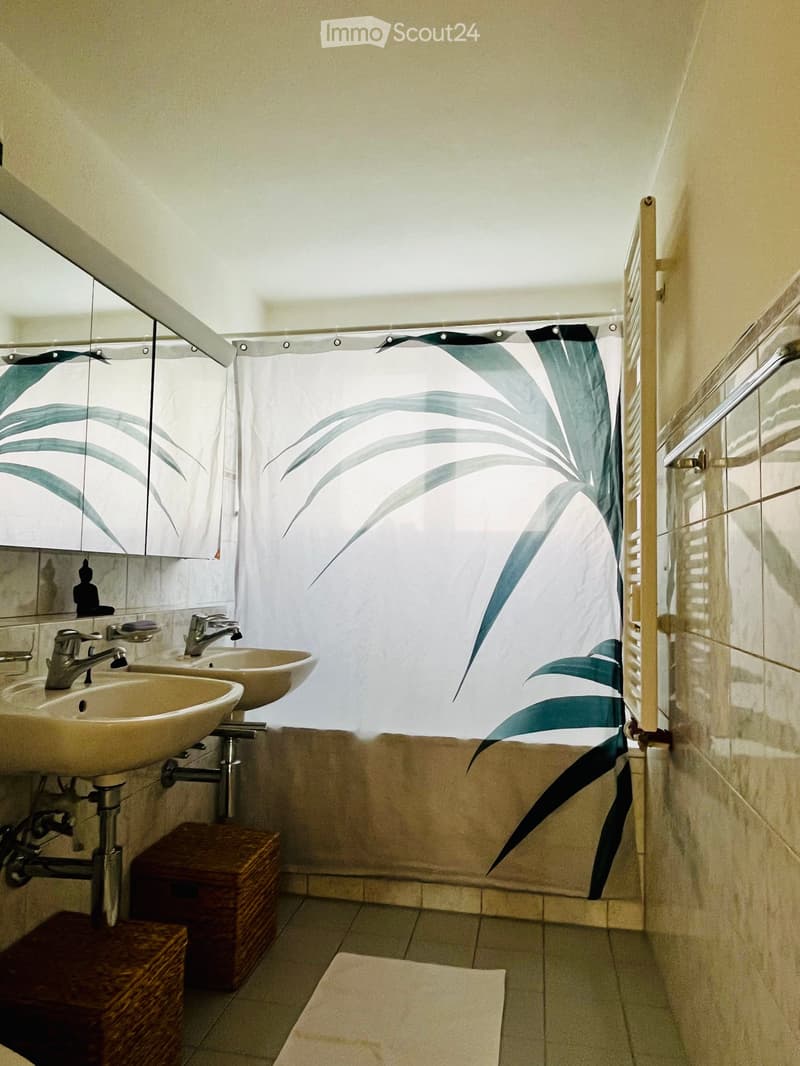 Modern bathroom with two sinks, a large mirror, a shower curtain with a leaf design, and a small wooden stool.