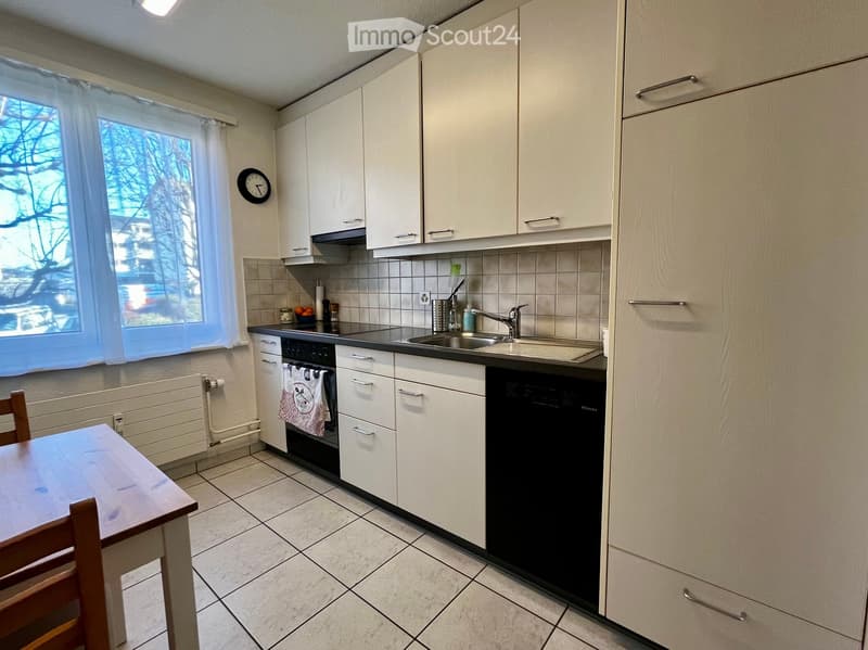 Modern kitchen with white cabinets, stainless steel sink, dishwasher, and a small dining area with a table and chair. The space features tiled flooring and a window with a view of trees.