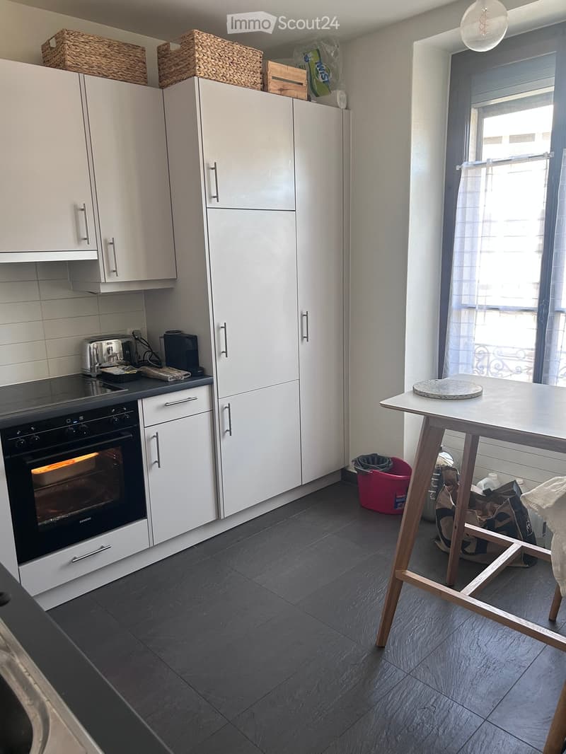 Modern kitchen with white cabinets, dark grey tiled floor, and a small wooden stool table near a window with a view.