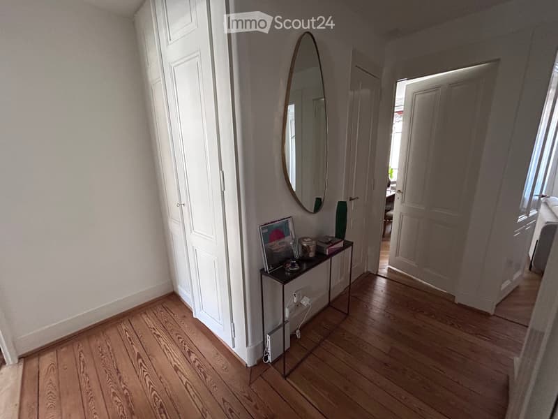 A narrow hallway with wooden flooring, white walls, and a small console table with a mirror and decorative items. Doors lead to other rooms.