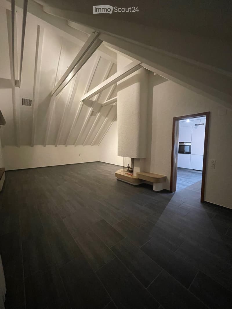 Empty attic space with wooden flooring, exposed wooden beams, and a small built-in bench near the wall. A doorway leads to another room with white cabinets and a built-in oven.