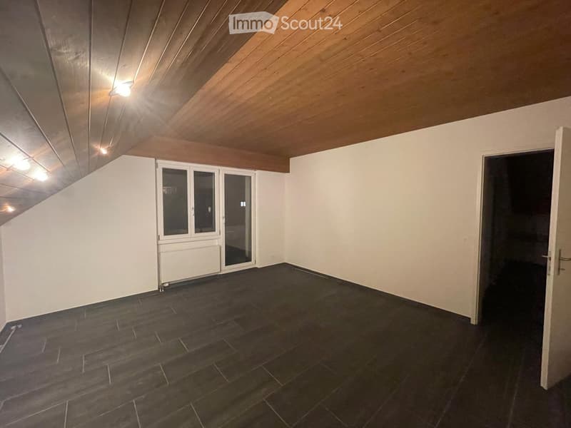 Empty room with dark tiled floor, white walls, wooden slanted ceiling, and a large window with glass doors leading outside.