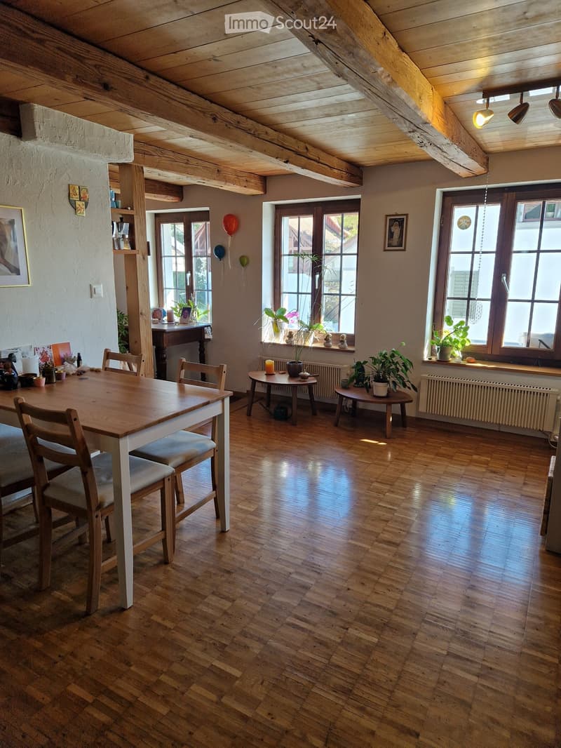 Bright, wooden-floored room with exposed wooden beams, large windows, and a mix of furniture including a dining table, chairs, small round tables, and potted plants. The room features a cozy, rustic design with warm lighting and decorative elements.