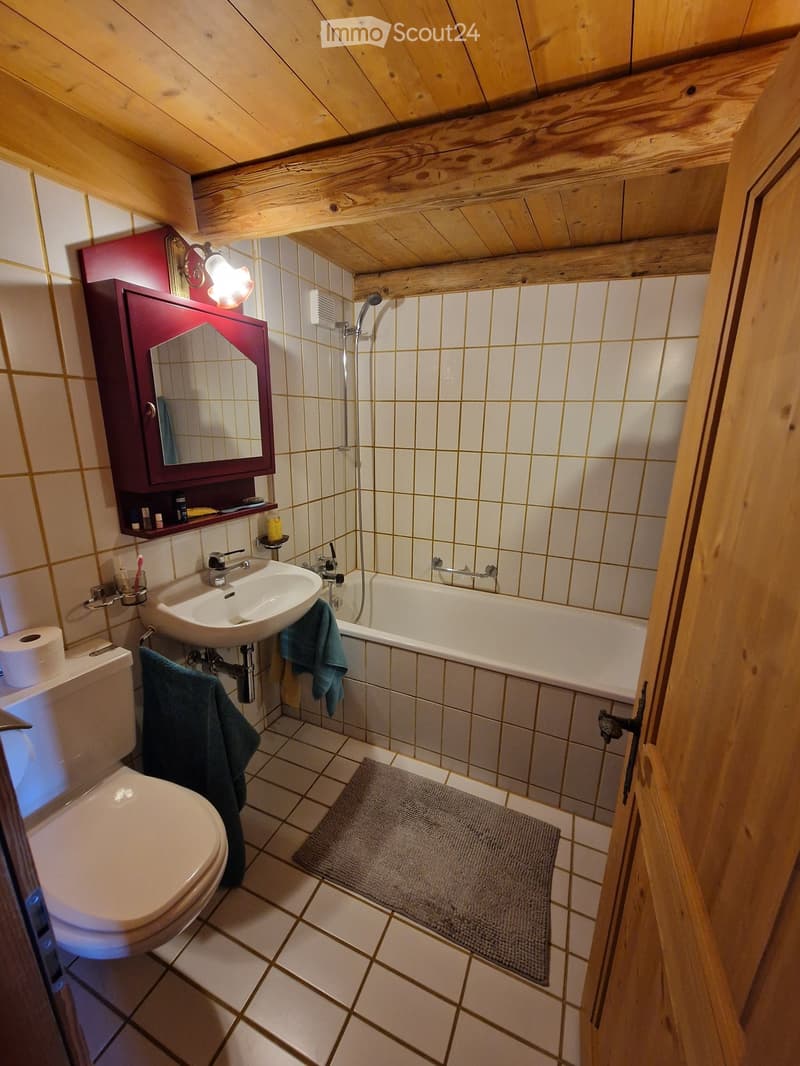 Cozy attic bathroom with wooden beams, white tiled walls, a toilet, sink, bathtub, and a small mirror cabinet. Towels are placed on the edge of the bathtub and sink.