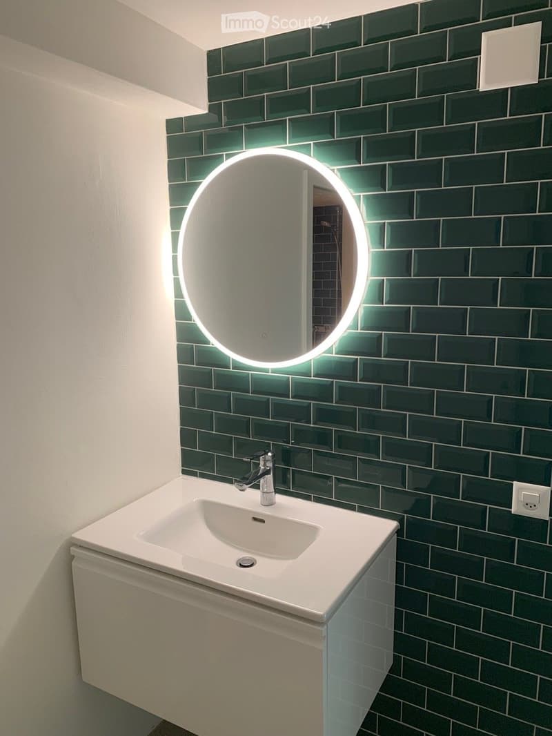 Modern bathroom with a white sink, chrome faucet, and a round illuminated mirror mounted on a dark green tiled wall.