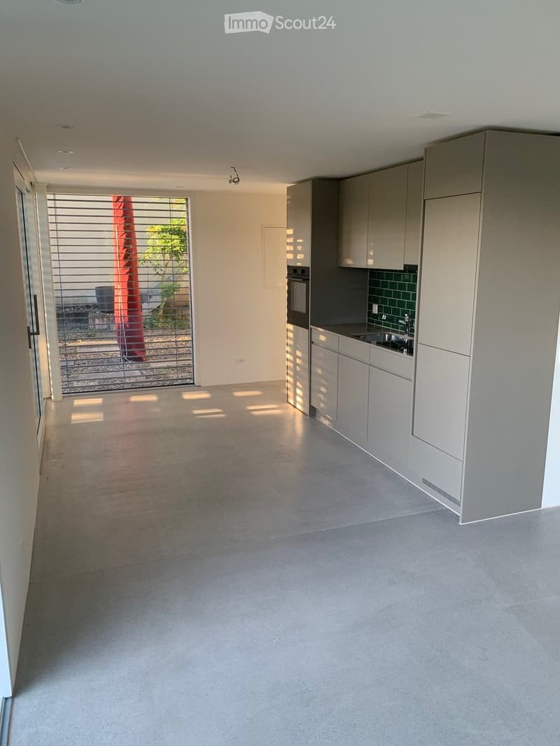Empty kitchen with modern grey cabinets, stainless steel appliances, and a large window with blinds leading to an outdoor area.