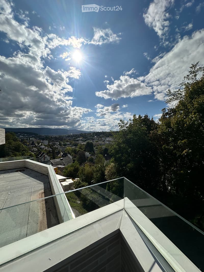 Glass-enclosed balcony with a panoramic view of a cityscape, trees, and distant hills under a partly cloudy sky.