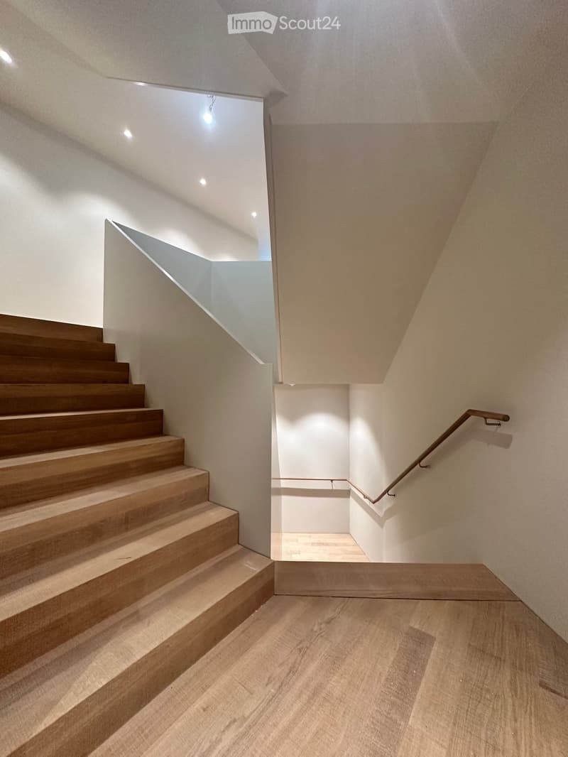 Modern staircase with wooden steps and a metal handrail, leading to an open area with recessed lighting and a view of another room.