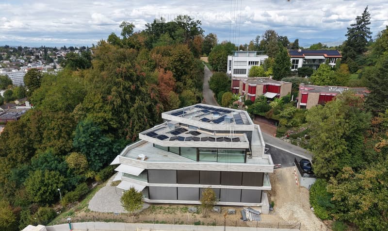 Modern two-story house with solar panels on the roof, surrounded by trees and greenery, with a parking area nearby.