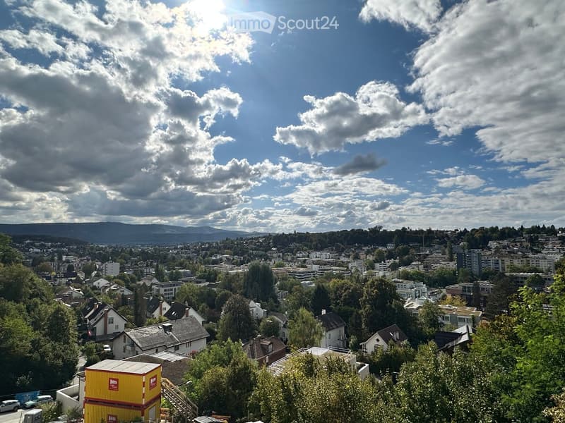 Panoramic view of a cityscape with residential buildings, trees, and distant mountains under a partly cloudy sky.