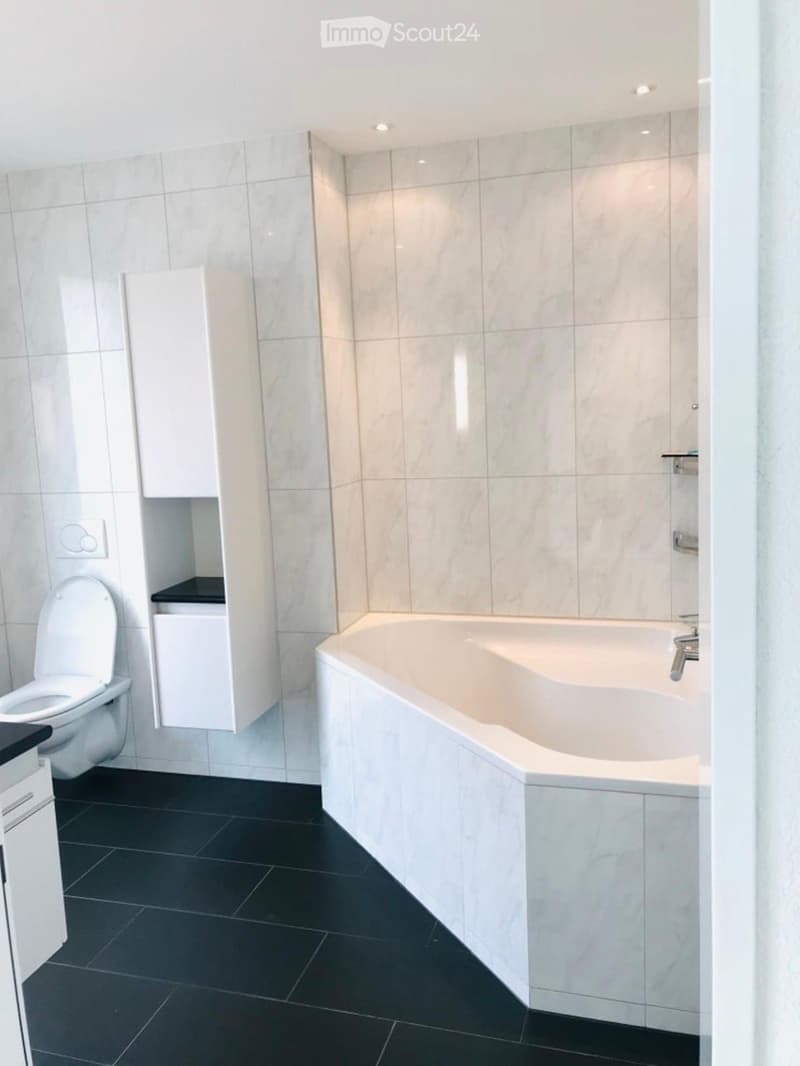 Modern bathroom with a white bathtub, toilet, and white cabinets. The walls are tiled in light marble, and the floor is black tile.