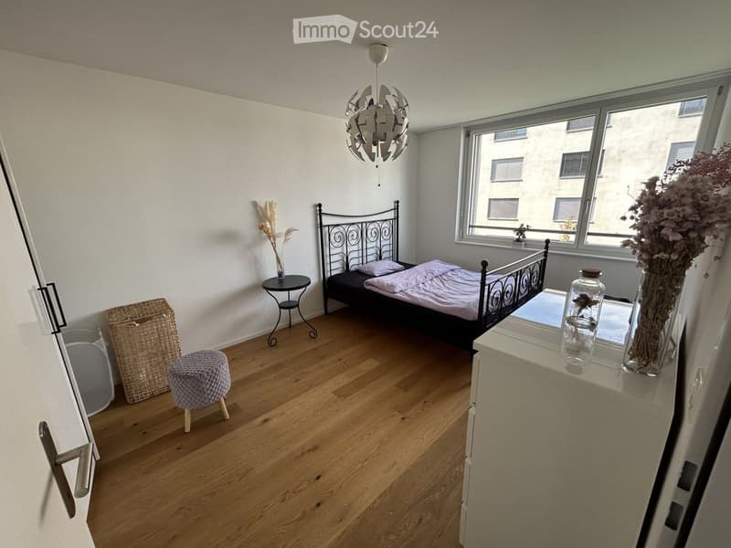 Cozy bedroom with wooden flooring, a metal-framed bed, a small round table, a wicker basket, a plush stool, and decorative vases on a nightstand.