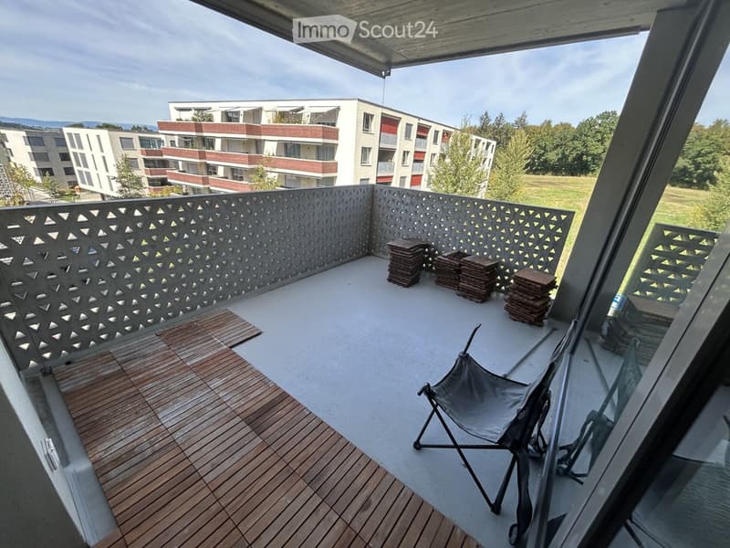 Balcony with wooden decking, grey folding chair, and stacks of wooden planks. Modern railing with geometric pattern.