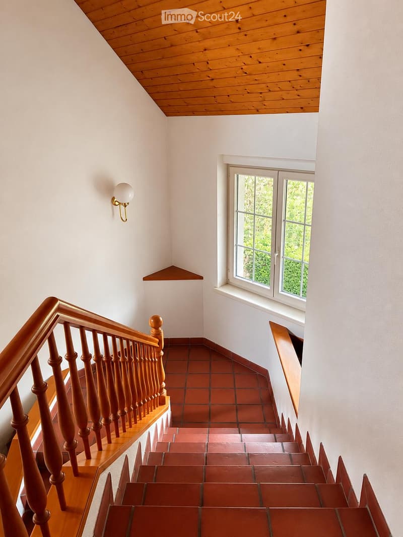 Spacious hallway with wooden staircase, red tiled steps, white walls, and a small window showing greenery outside.