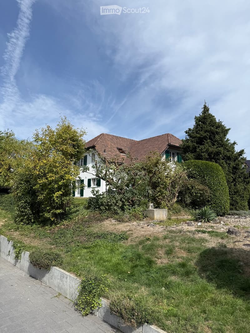 Two-story house with a red-tiled roof, white walls, green shutters, surrounded by trees and shrubs, with a raised garden area in front.