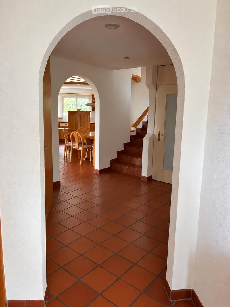 A spacious hallway with terracotta tiled flooring and white walls, featuring an arched doorway leading to a dining area with wooden furniture and a staircase leading upwards.