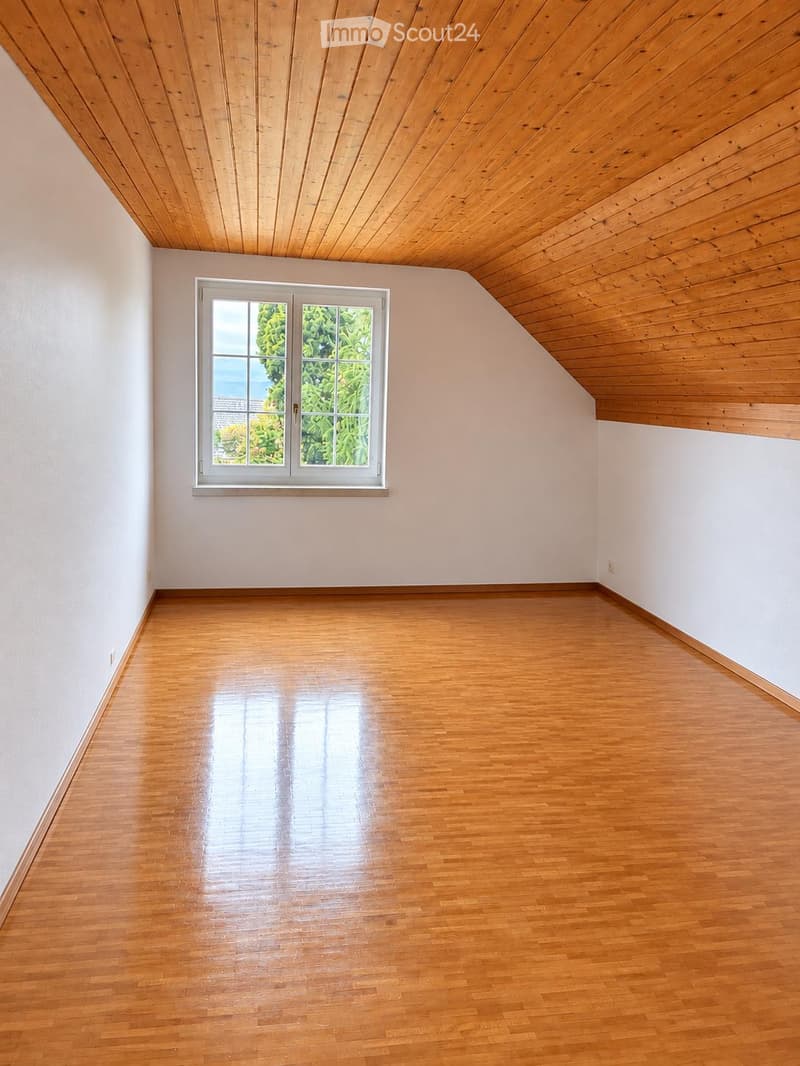 Empty room with wooden flooring and white walls, featuring a large window with a view of greenery outside.