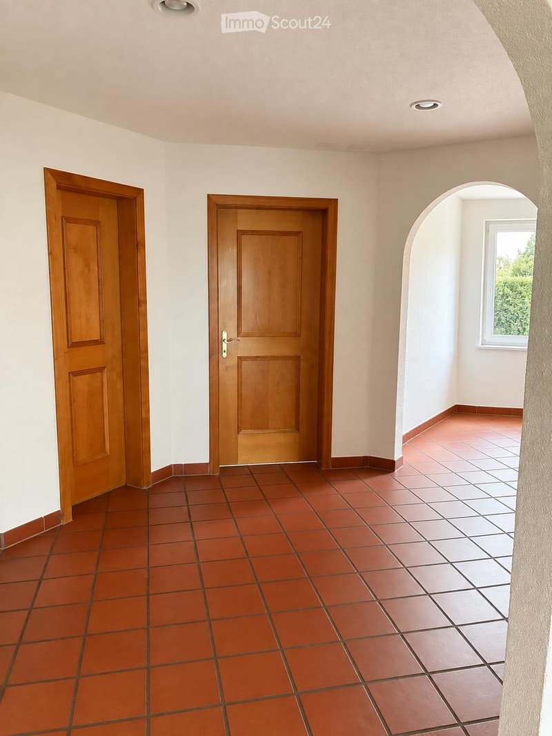 Empty hallway with red tiled flooring, white walls, and two wooden doors. An arched opening leads to a room with a window and additional tiles.