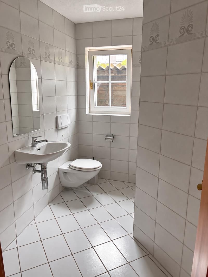 A small bathroom with white tiled walls and floor, featuring a sink with a modern faucet, a toilet, a mirror, and a window offering a view of the roof.