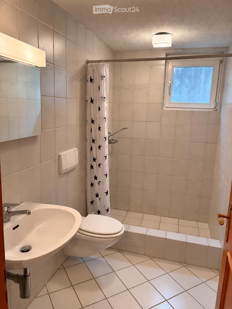 Bathroom with white tiled walls and floor, a sink, toilet, shower curtain with star pattern, and a small window.