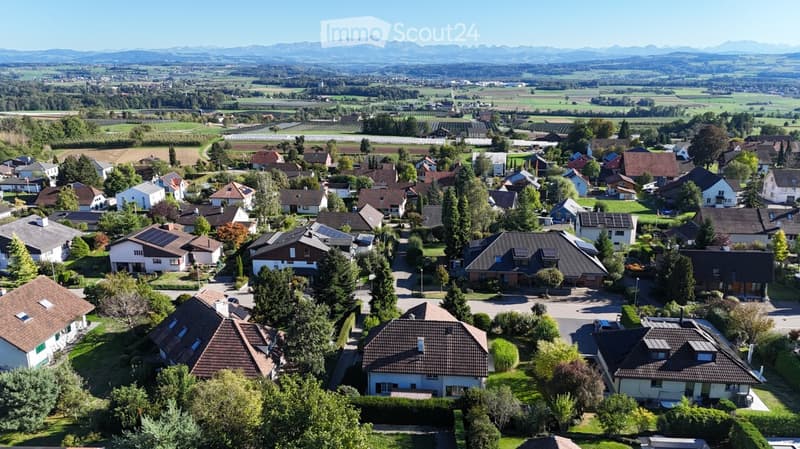 Aerial view of a suburban neighborhood with numerous houses, surrounded by green fields and distant mountains.