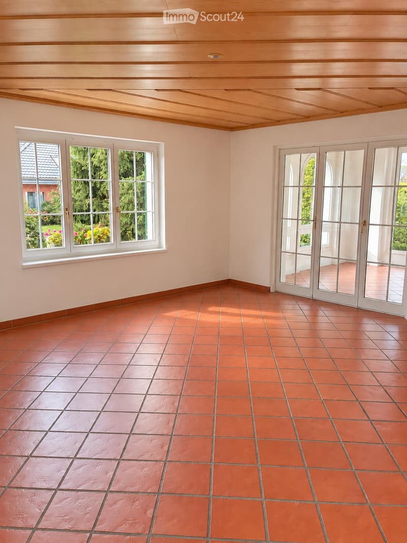 Empty room with terracotta tiled floor, white walls, large windows, and sliding glass doors leading outside.