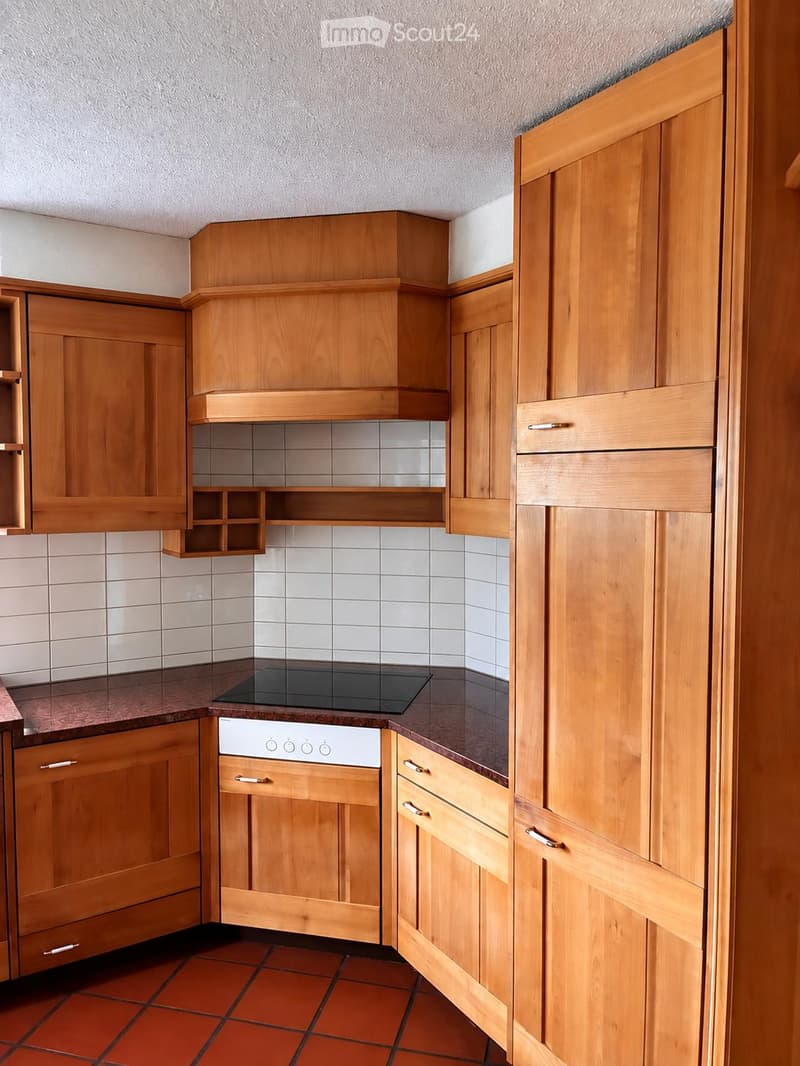 Modern kitchen with wooden cabinets, electric stove, oven, and kitchen island. The countertops are dark brown, and the floor is tiled in a warm orange color.