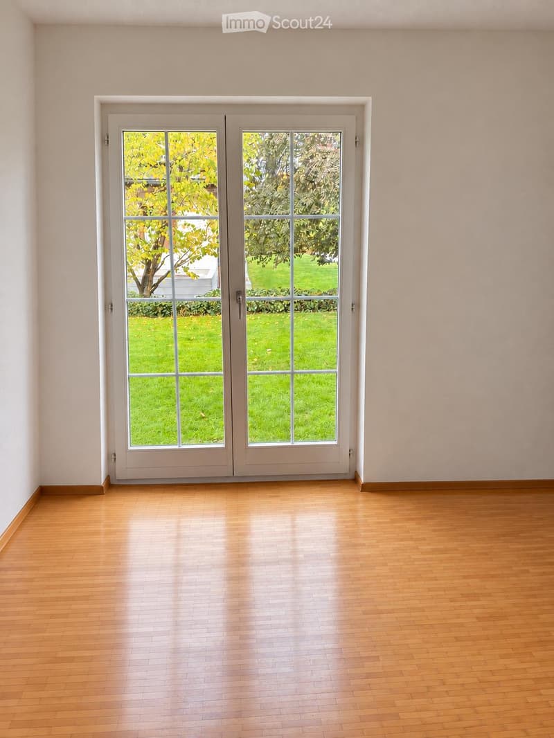 Empty room with wooden flooring and large double doors leading to a green outdoor area.