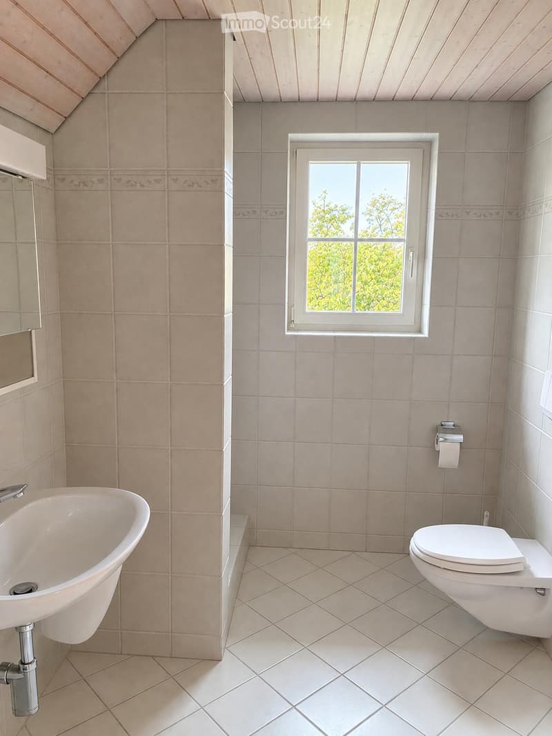 Bathroom with beige tiles, a bathtub, toilet, and a window with a view of greenery.