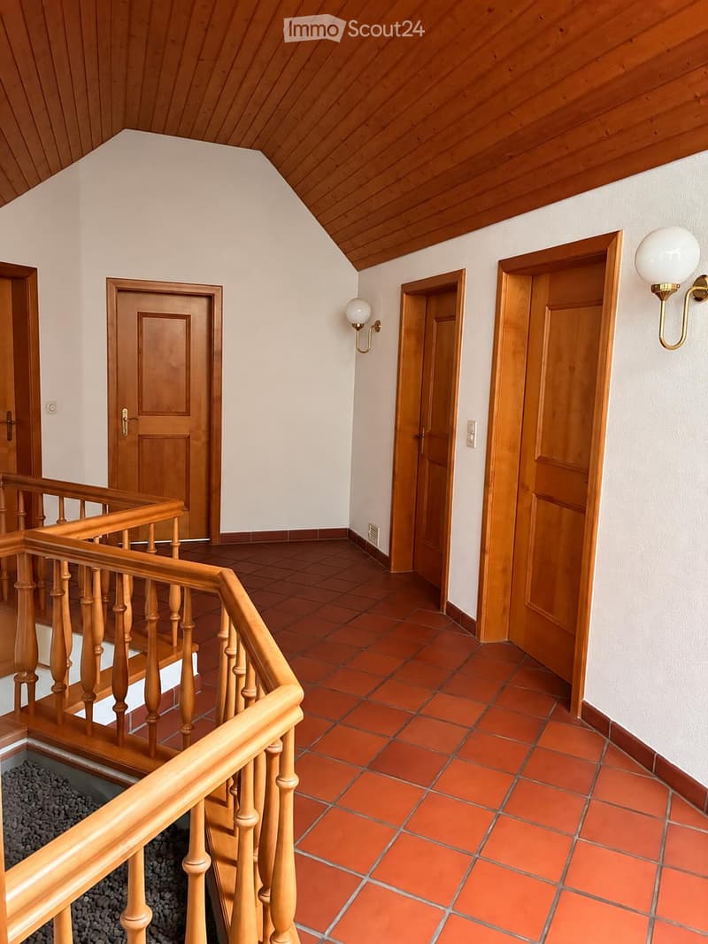 Wooden staircase with a wooden railing, red tiled floor, white walls, and several wooden doors leading to different rooms.