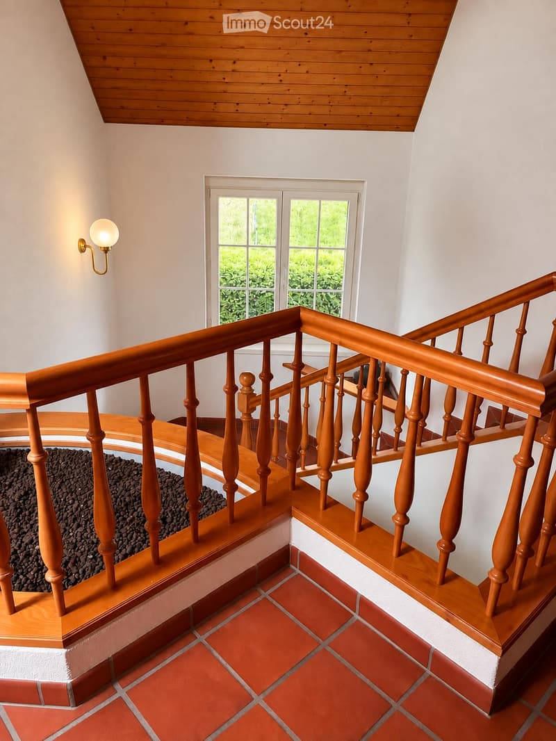 Spacious hallway with wooden staircase, red-tiled floor, white walls, and a window overlooking greenery.