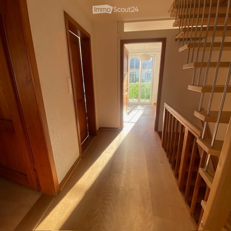 Bright hallway with wooden flooring, leading to a staircase and an open doorway to a room with large windows and a view of greenery.