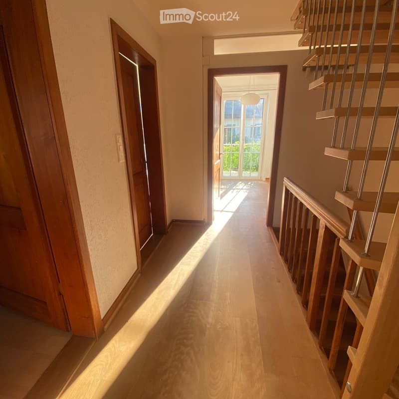 Bright hallway with wooden flooring, modern staircase with metal railings, and open doors leading to other rooms.