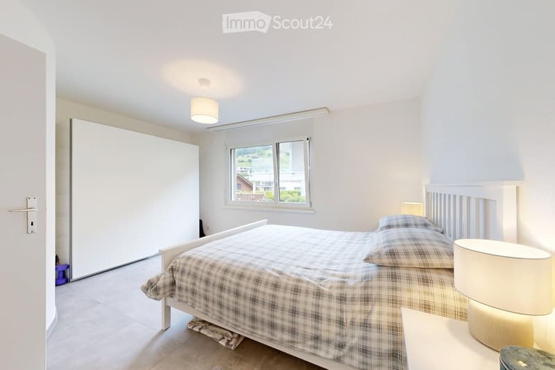 Bright bedroom with a double bed, plaid duvet, nightstand with lamp, and a large window with blinds. The room features a white wardrobe and a modern ceiling light.