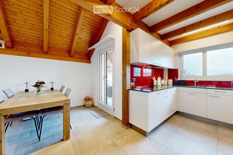 Bright, open-plan room with a wooden dining table and chairs, a modern kitchen area with white cabinets and a red backsplash, and large windows offering natural light.