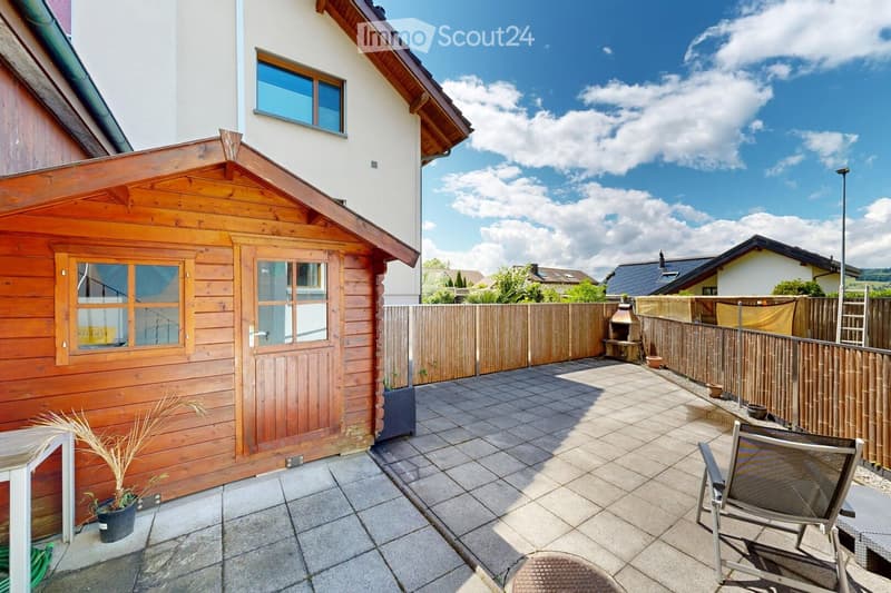 A tiled patio area with a wooden shed, potted plants, a barbecue, and a lounge chair. The area is enclosed by a wooden fence and features a clear blue sky with scattered clouds in the background.