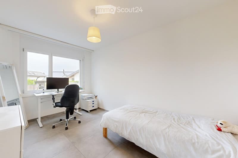 Bright bedroom with a bed, desk, chair, computer, and a large window offering a view of the outside. The room features a modern, minimalist design with white walls and a tiled floor.