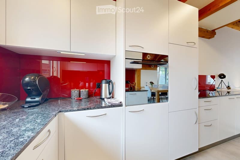 Modern kitchen with white cabinets, granite countertop, coffee maker, kettle, and a small dining area visible through an open doorway.