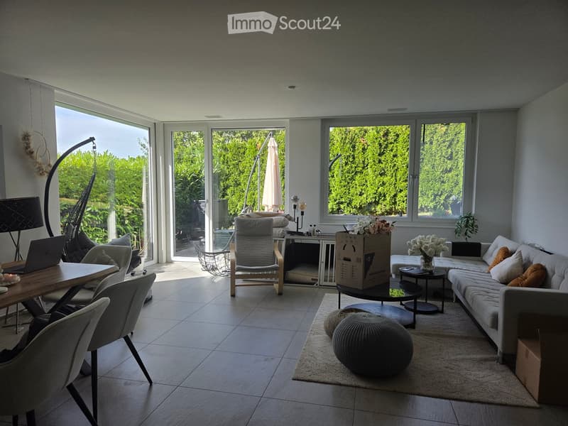 Bright living room with large windows and sliding doors leading to a patio, featuring a grey sofa, coffee table, round stools, dining table with chairs, and decorative vases with flowers.