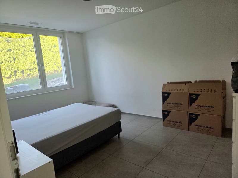 Bedroom with a bed, nightstand, and several cardboard boxes stacked against the wall. Large windows provide natural light.