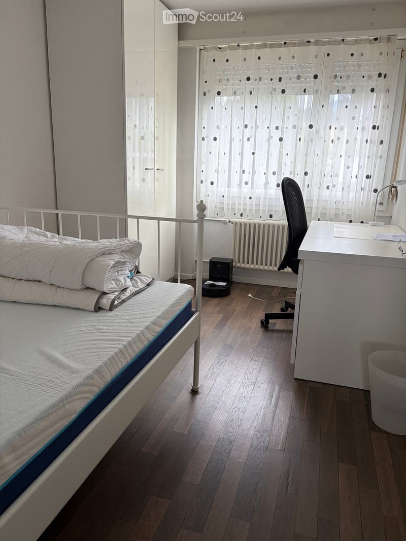 A tidy bedroom with a single bed, desk, chair, radiator, and a window with curtains. The room features wooden flooring and a white wardrobe.